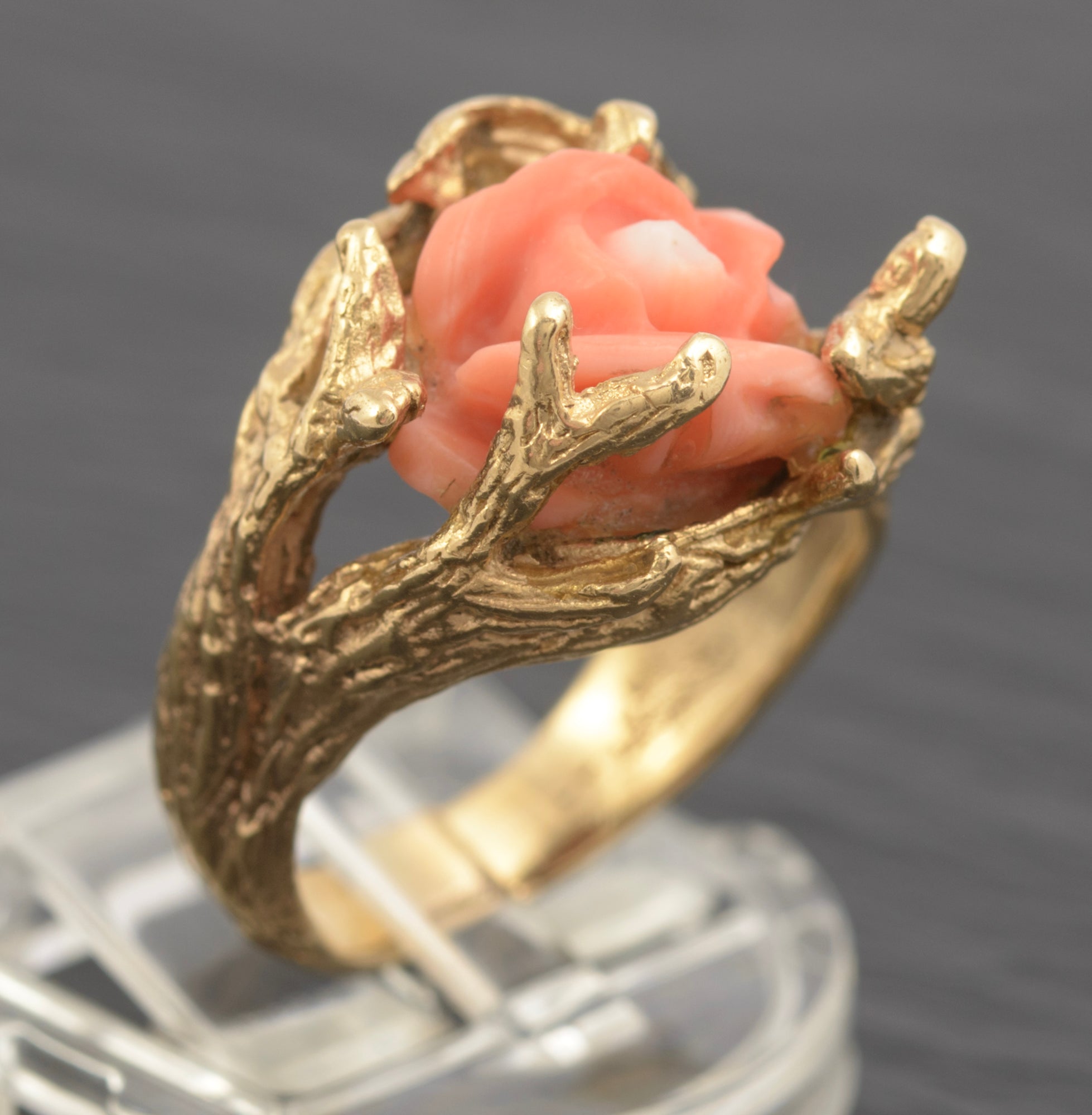 A gold ring featuring a carved pink coral rose nestled within branch-like gold settings, displayed on a clear stand against a dark gray background.
