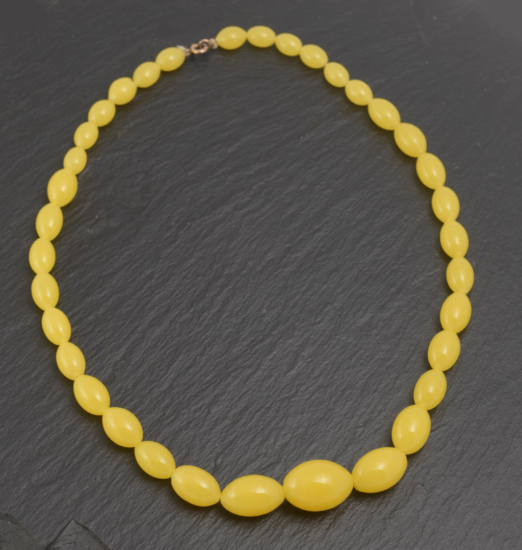 Art Deco uranium vaseline glass bead necklace - vivid opaque yellow - full view laid flat on dark slate showing graduating oval beads from small clasp end to large central oval bead