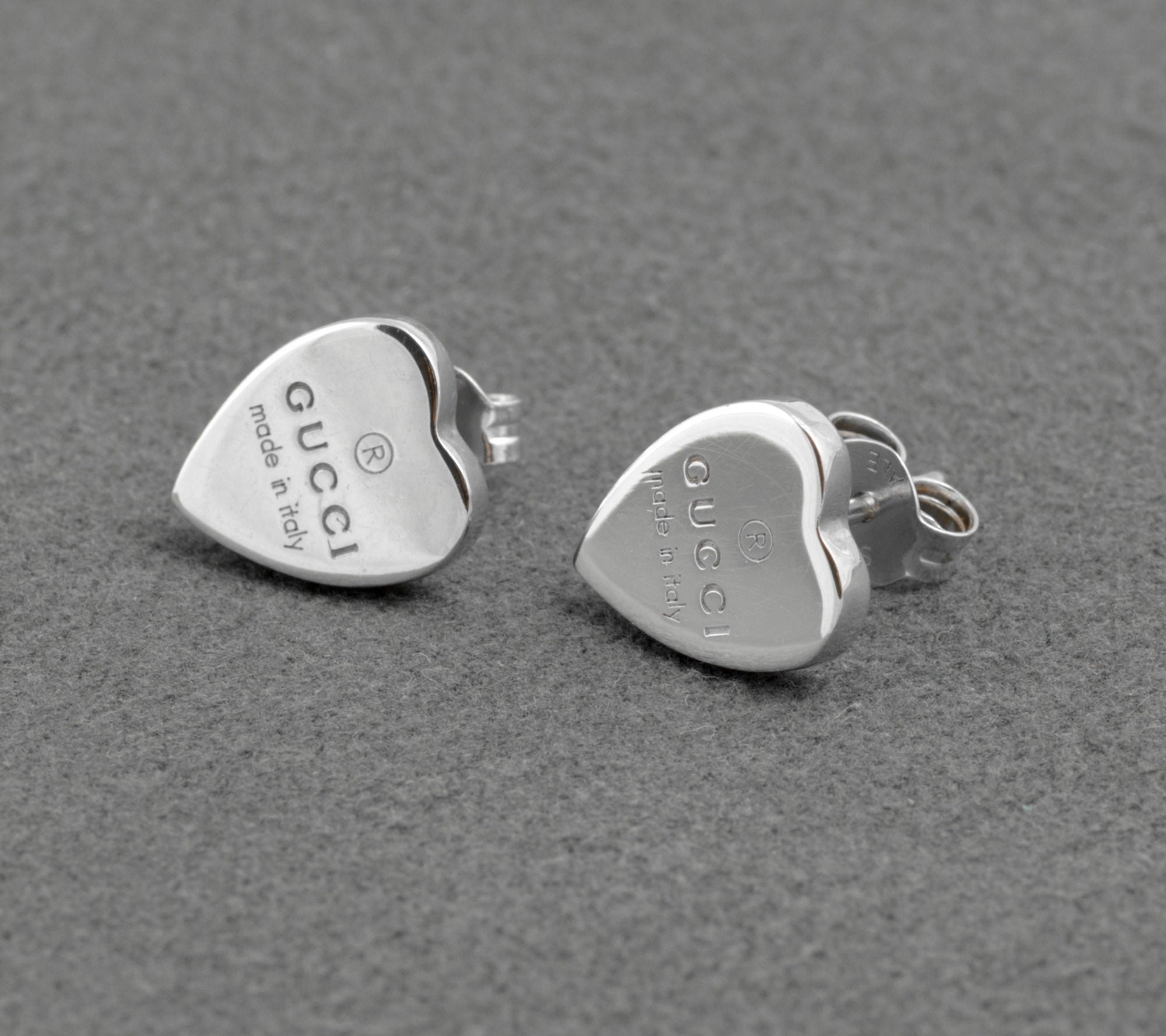 Gucci sterling silver heart stud earrings - made in Italy - pair shown from front and side on grey background showing polished heart form with Gucci name and made in italy engraving and butterfly back fittings