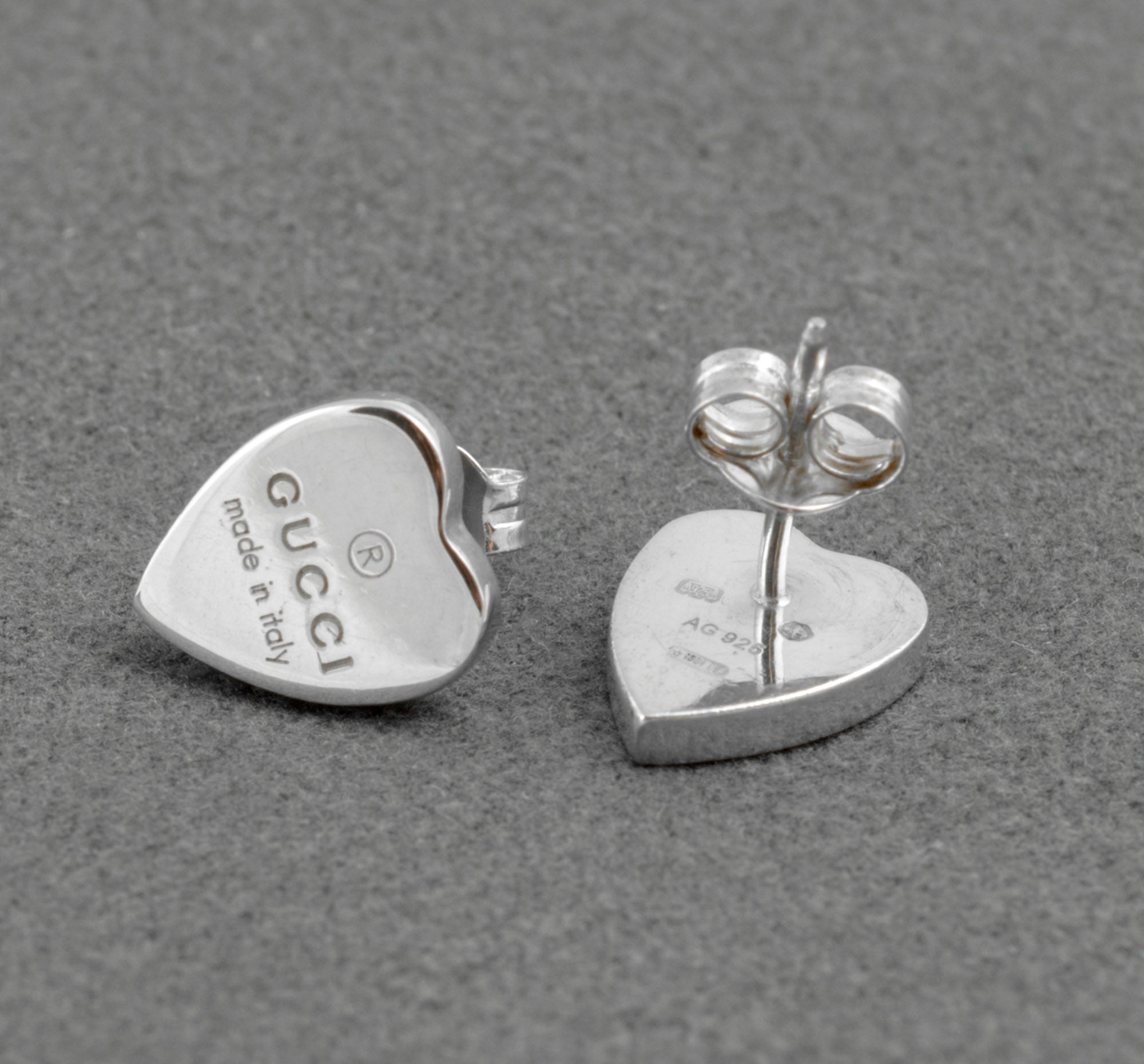 Gucci sterling silver heart stud earrings - made in Italy - one earring front showing Gucci engraving, one earring reverse showing AG 925 hallmark and Florence assay mark with post and butterfly back separated on grey background