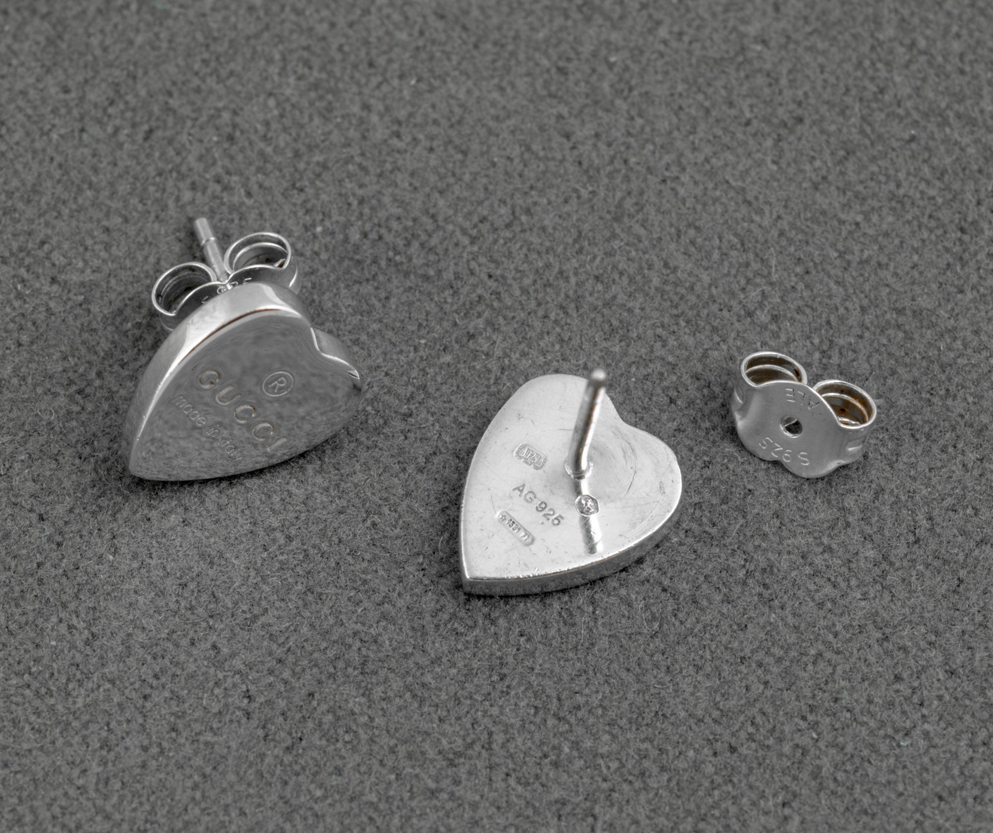 Gucci sterling silver heart stud earrings - made in Italy - three piece detail showing earring front with Gucci engraving, earring reverse with AG 925 mark, and butterfly back fitting separately on grey background