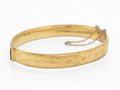 Vintage 1/5 9ct rolled gold hinged bangle with hand engraved floral and scrolling foliage decoration and rope twist border edges three quarter view showing clasp and safety chain on white background