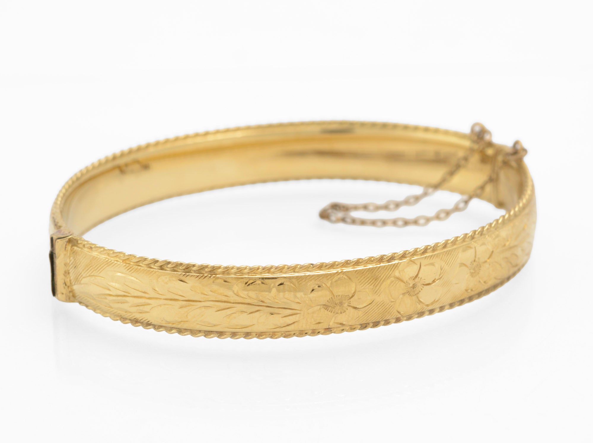 Vintage 1/5 9ct rolled gold hinged bangle with hand engraved floral and scrolling foliage decoration and rope twist border edges three quarter view showing clasp and safety chain on white background