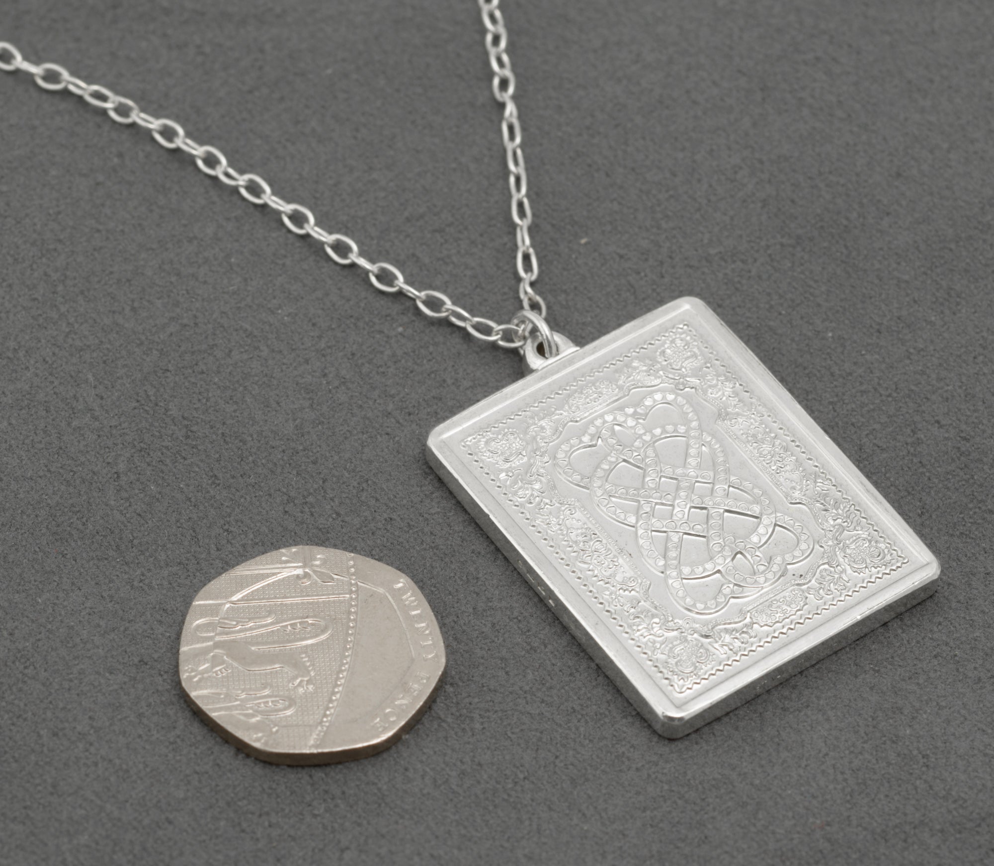 Vintage sterling silver John Pinches 1977 St Valentine's Day pendant with silver chain laid flat on grey background next to 20p coin showing scale of pendant against coin