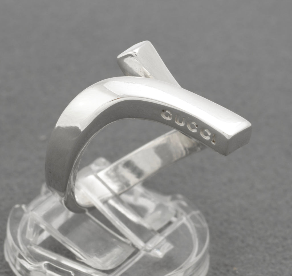 Gucci sterling silver crossover ring on clear ring stand showing crossover structure with upper diagonal branded bar and lower squared end with GUCCI lettering visible on grey background