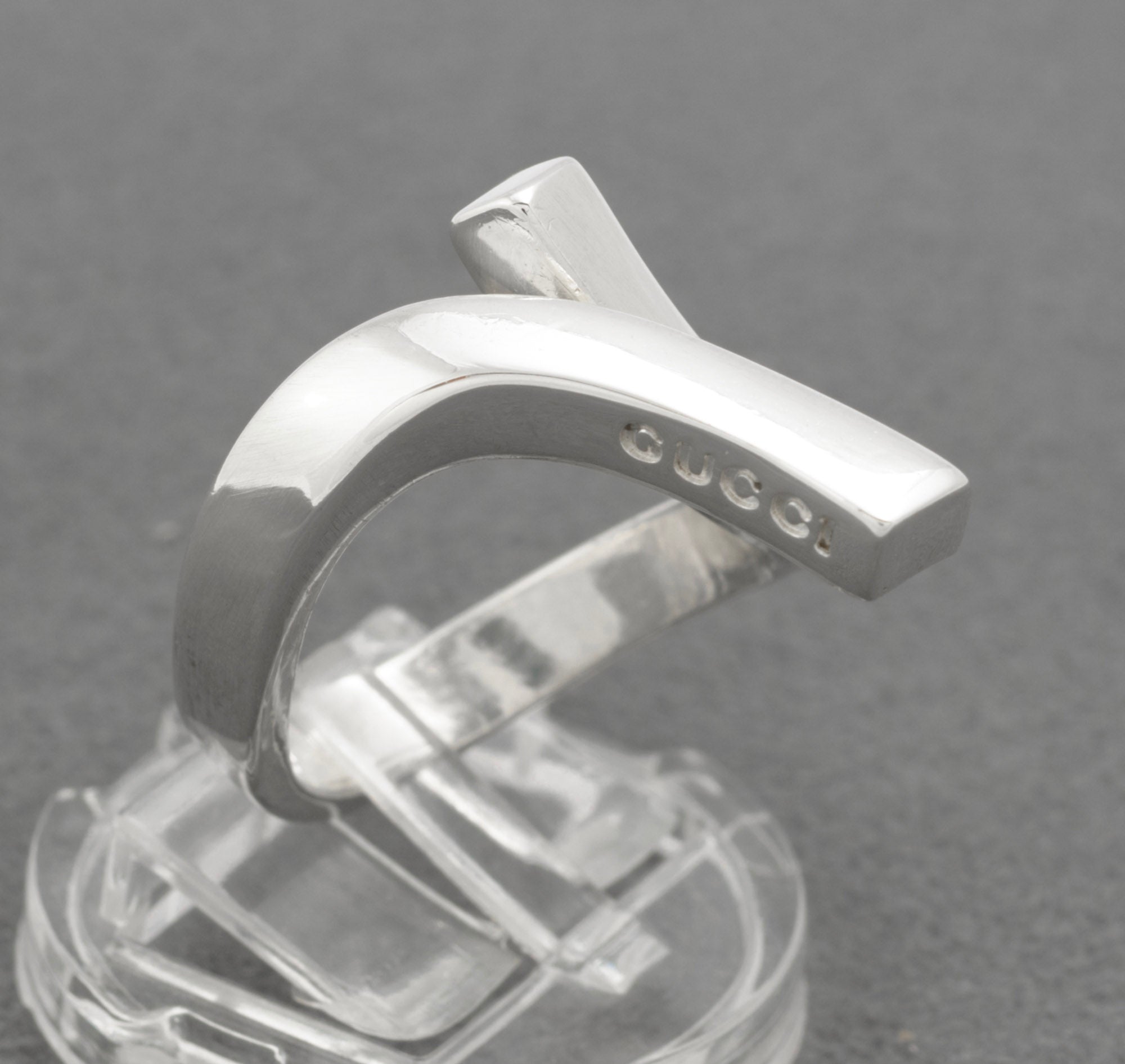 Gucci sterling silver crossover ring on clear ring stand showing crossover structure with upper diagonal branded bar and lower squared end with GUCCI lettering visible on grey background
