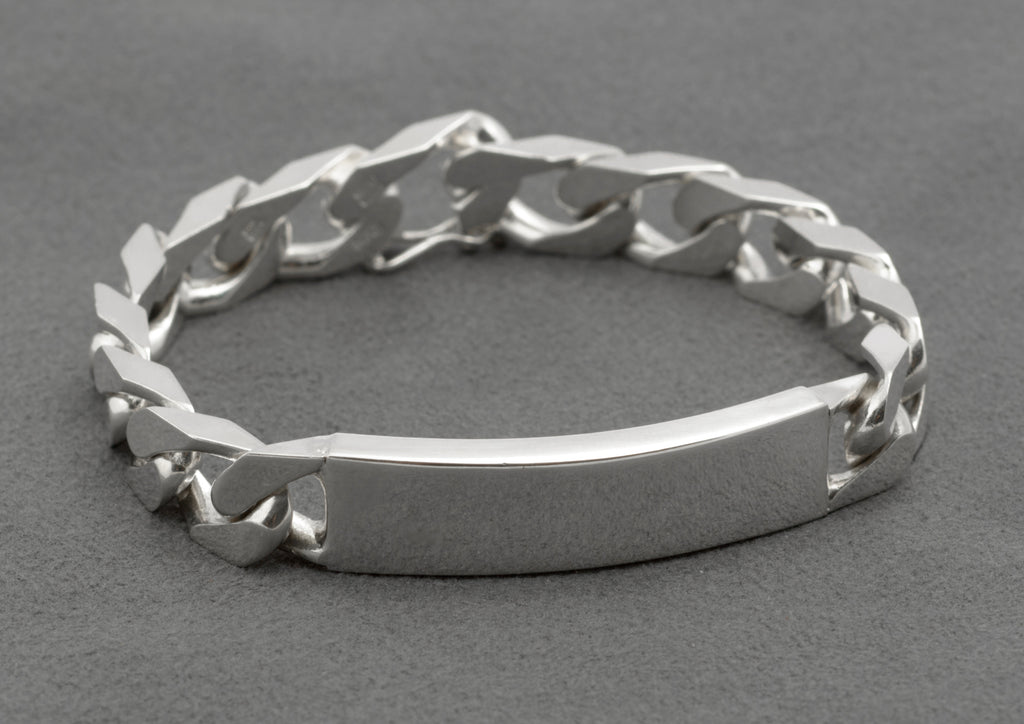 Italian sterling silver ID bracelet with broad flattened curb links and plain curved ID panel laid in circle on dark grey background showing full construction and push-piece box clasp