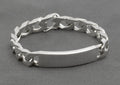 Italian sterling silver ID bracelet with broad flattened curb links and plain curved ID panel laid in circle on dark grey background showing full construction and push-piece box clasp