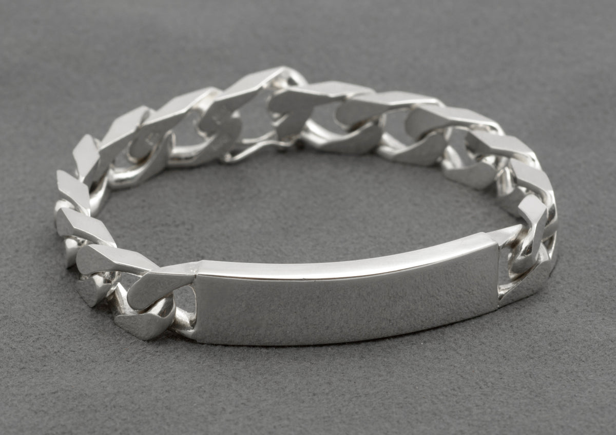 Italian sterling silver ID bracelet with broad flattened curb links and plain curved ID panel laid in circle on dark grey background showing full construction and push-piece box clasp