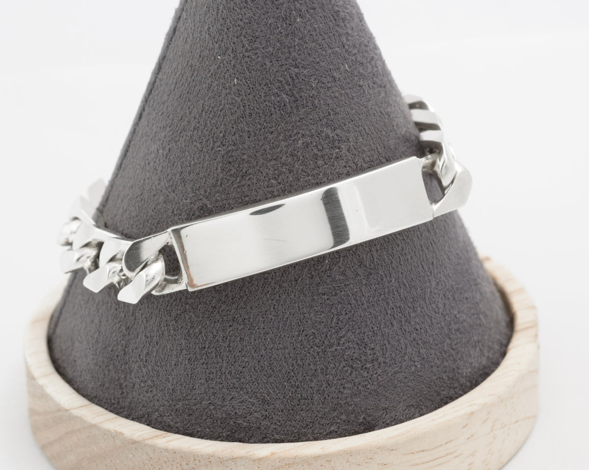 Italian sterling silver ID bracelet laid flat on light grey background showing full length plain curved ID panel and broad flattened curb links with integrated push-piece box clasp