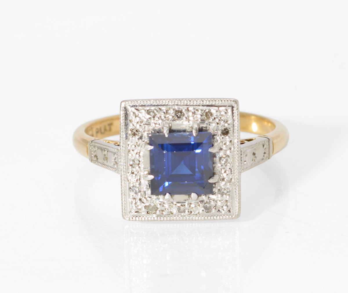An Art Deco period ring, circa 1930, made in 18ct yellow gold with a platinum head and upper shoulders. Set with a vivid royal blue square cut sapphire surrounded by twelve small diamonds, with further illusion style detailing to the shoulders. Marked 18CT PLAT to the inside of the band. UK size N. Supplied with the period jewellery box shown.
