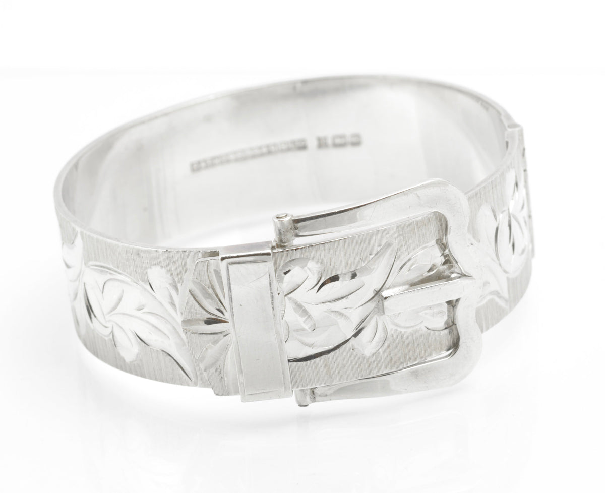 A vintage sterling silver cuff bangle by Bracelon, hallmarked Birmingham 1976, fitted with an unusual patented buckle fastening. The broad textured band is decorated with bright cut scrolling foliate decoration, and the buckle face carries matching engraved detail. 49.6 grams.