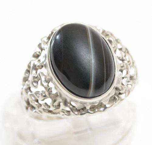 A silver ring featuring a large oval black stone with fine white banding, displayed on a clear stand against a white background.